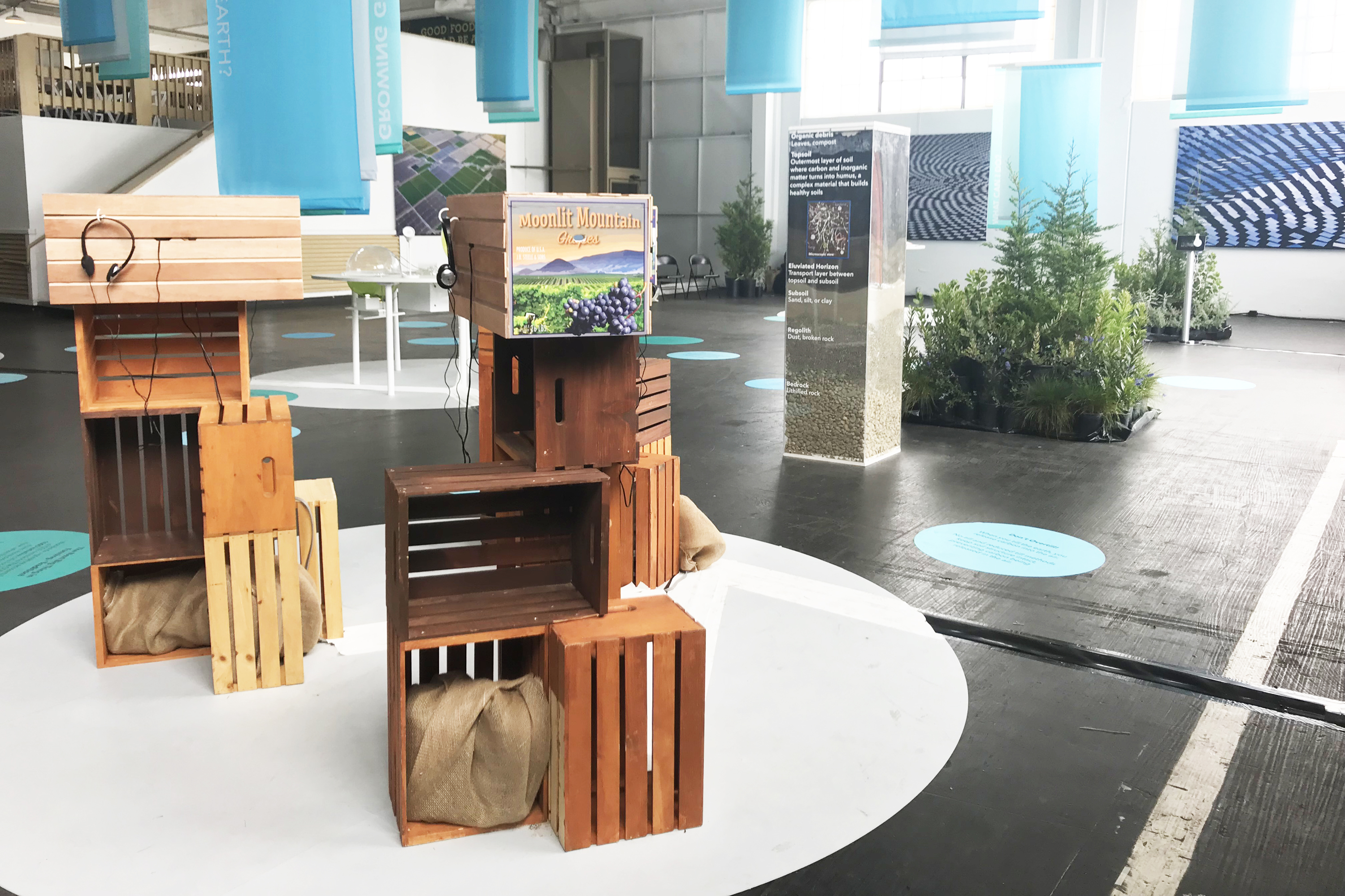 Three fruit crates with brand graphics, a small peephole, and headphones on top of several other smaller fruit crates, with a large soil sculpture in the background
